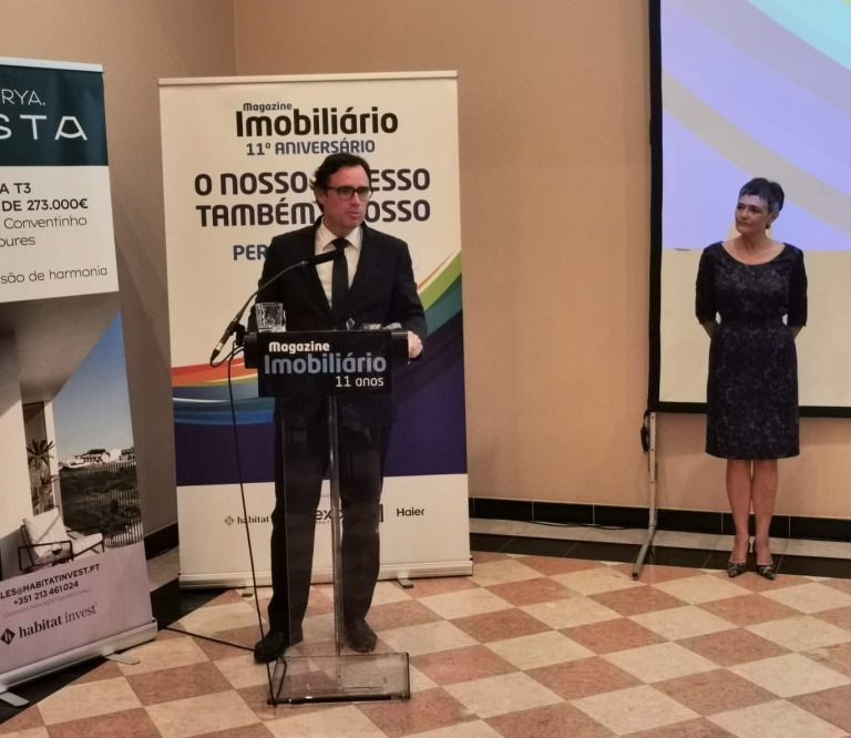 Nelson Lage está de fato escuro e óculos, fala ao microfone num púlpito transparente, durante evento comemorativo do 11.º aniversário da revista Magazine Imobiliário. Ao fundo, um cartaz com o lema "O nosso sucesso também é vosso". À direita da imagem, uma mulher de vestido escuro observa com um sorriso discreto, junto a um ecrã com projecção gráfica.