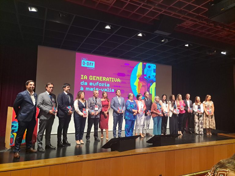 Grupo de cerca de vinte pessoas está alinhado no palco de um auditório, durante o evento Q-Day Conference 2024. Ao fundo, um ecrã exibe o tema “IA Generativa: da euforia à mais-valia”, com grafismo colorido em tons de rosa, roxo e azul. Os participantes seguram troféus ou lembranças, posando para fotografia oficial num momento de distinção.