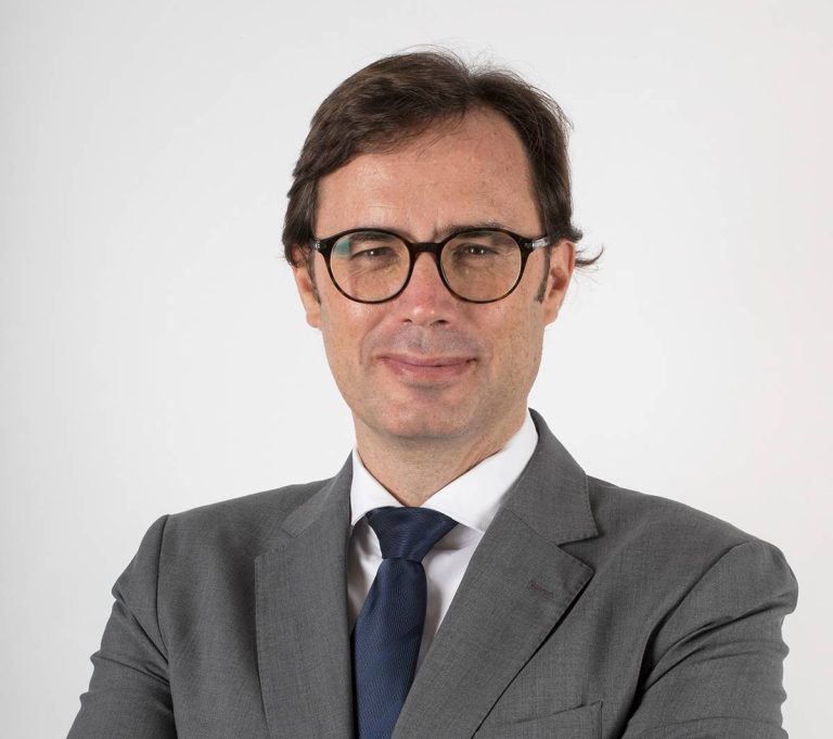 Retrato profissional do presidente da Adene. Um homem de cabelo castanho curto, a usar óculos redondos de aro escuro, fato cinzento e gravata azul-escura. Está de braços cruzados e sorri levemente, com fundo branco neutro.