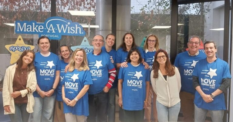 Grupo de 13 pessoas posa sorridente em frente a uma janela com o logótipo “Make-A-Wish Portugal”. A maioria veste t-shirts azuis com a frase “MOVE FOR WISH KIDS” e uma estrela branca no centro, indicando participação numa acção solidária. Duas pessoas usam roupa casual sem a t-shirt do evento.