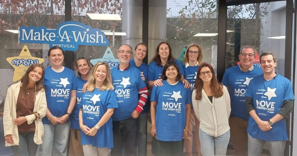 Grupo de 13 pessoas posa sorridente em frente a uma janela com o logótipo “Make-A-Wish Portugal”. A maioria veste t-shirts azuis com a frase “MOVE FOR WISH KIDS” e uma estrela branca no centro, indicando participação numa acção solidária. Duas pessoas usam roupa casual sem a t-shirt do evento.