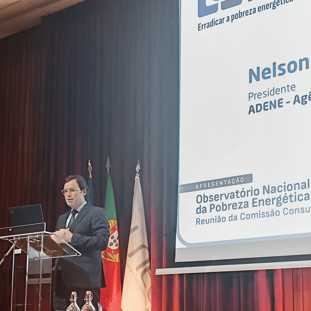 Nelson Lage de fato escuro e óculos fala num púlpito de acrílico em frente a um ecrã de projecção que exibe o título “Observatório Nacional da Pobreza Energética – Reunião da Comissão Consultiva”. À esquerda do orador estão as bandeiras de Portugal, da União Europeia e da ADENE. O evento decorre num auditório com cortinas vermelhas.