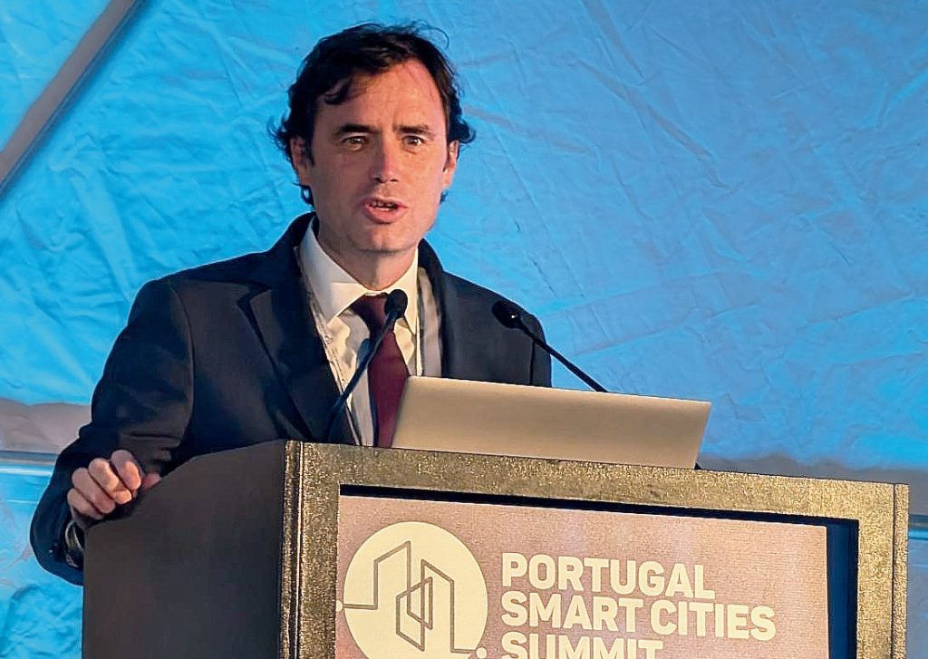 Nelson Lage de fato escuro e gravata bordô fala ao microfone num púlpito durante o evento Portugal Smart Cities Summit. À frente do púlpito está o logótipo do evento com o nome em letras brancas. O fundo azul-claro indica um espaço de evento coberto.