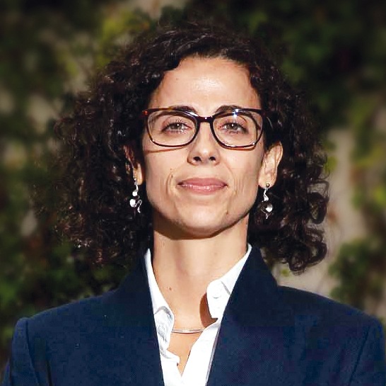 Retrato formal de Marina Alves. Uma mulher com cabelo castanho escuro, encaracolado e solto, a usar óculos de armação escura, brincos compridos e um blazer azul-escuro sobre camisa branca. Está com expressão serena, fotografada em ambiente exterior com fundo desfocado de vegetação.