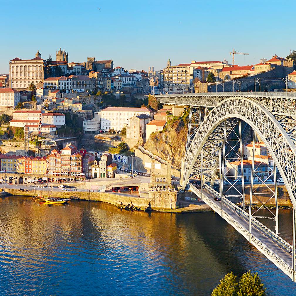 Vista panorâmica da cidade do Porto ao final da tarde, com destaque para a ponte metálica Luís I sobre o rio Douro, iluminada pela luz dourada do sol. À esquerda, vê-se o casario tradicional da Ribeira e, ao fundo, edifícios históricos como a Sé do Porto.