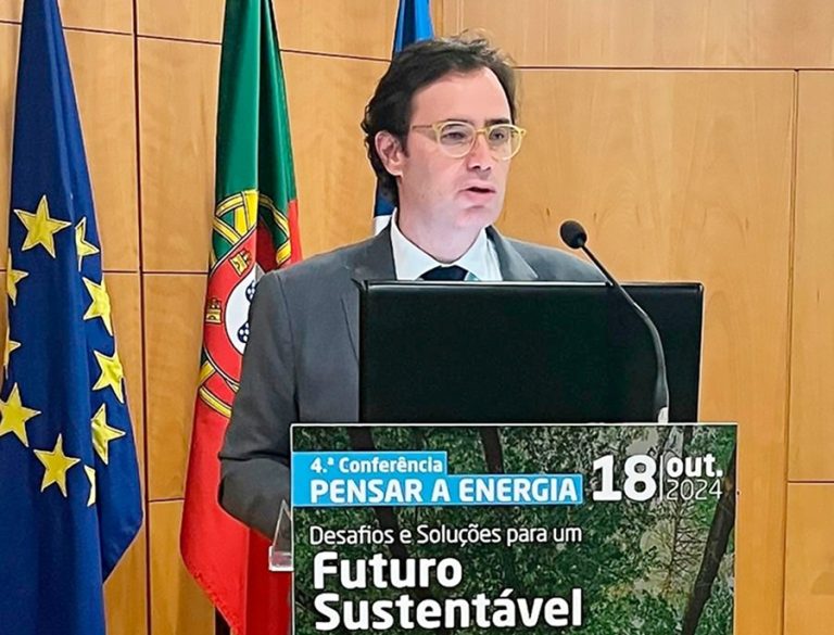 Nelson Lage com fato escuro e óculos fala ao púlpito durante a 4.ª Conferência Pensar a Energia. À frente do púlpito, há um cartaz do evento com os dizeres: “Desafios e Soluções para um Futuro Sustentável – 18 out. 2024”. Ao fundo, estão visíveis as bandeiras da União Europeia, de Portugal e da Universidade do Algarve.