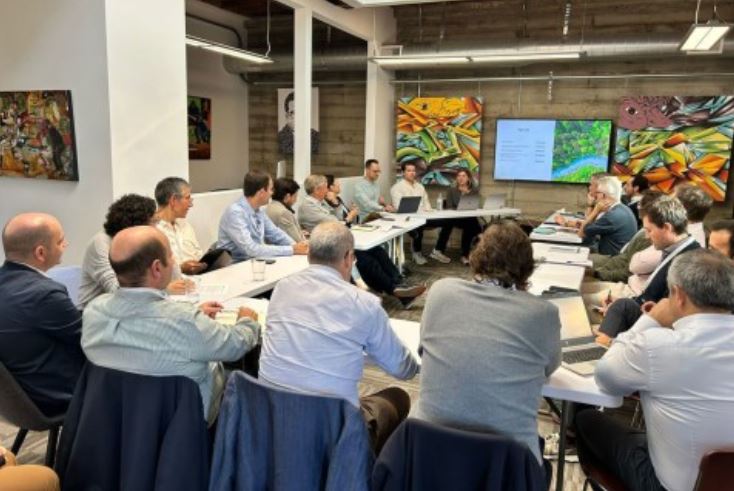 Cerca de 18 pessoas estão reunidas em torno de uma mesa em formato U, participando numa reunião do GSI Energy. A sala é moderna, com paredes de madeira e quadros coloridos. Ao fundo, um ecrã exibe uma apresentação com o título “GSI Energy” e imagens de natureza, sugerindo temas ligados à sustentabilidade ou energia.