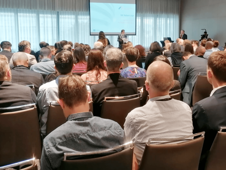 Fotografia de um auditório cheio de participantes, vistos de costas, todos sentados e atentos a uma apresentação. Ao fundo, vê-se um palco com uma oradora a falar ao púlpito. No ecrã atrás dela aparece o nome "Mr. Nelson Lage". A sala é moderna, com luz natural a entrar por grandes janelas cobertas com cortinas translúcidas. Há também um operador de câmara a registar o evento.