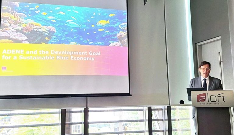 Fotografia de uma apresentação no congresso "Ocean 2024", em Singapura. O orador, em fato e gravata, encontra-se ao púlpito com o logótipo "Aloft Singapore Novena". No ecrã é visível o slide com o título: “ADENE and the Development Goal for a Sustainable Blue Economy”, acompanhado por uma imagem colorida de um recife de coral com peixes tropicais.