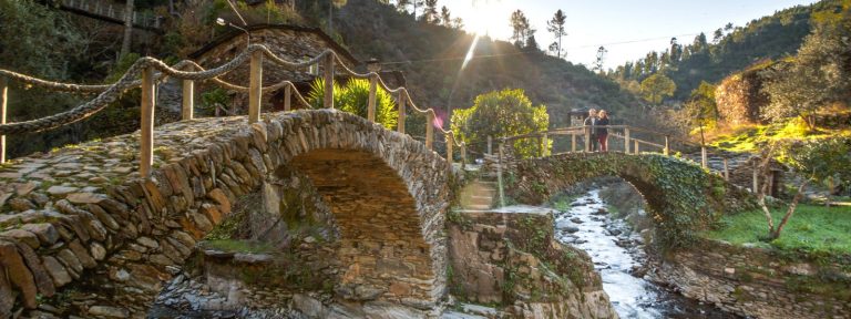 Dois pontes de pedra rústica atravessam um pequeno riacho ladeado por vegetação verdejante, numa paisagem montanhosa iluminada pela luz dourada do sol poente. Ao fundo, vêem-se casas construídas em pedra e duas pessoas a observar a paisagem a partir da ponte.