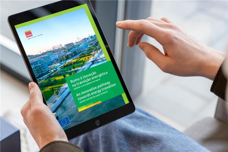 Uma pessoa segura um tablet com as duas mãos, onde é visível a capa de uma publicação digital da ADENE. A capa tem uma fotografia aérea de uma zona urbana moderna, com espaços verdes, edifícios e painéis solares. O título da publicação é “Rumo à inovação na transição energética – ADENE e os projectos europeus” e o subtítulo em inglês diz: “An innovative pathway towards energy transition – ADENE and the European projects”. Estão presentes os logótipos da ADENE e da União Europeia.