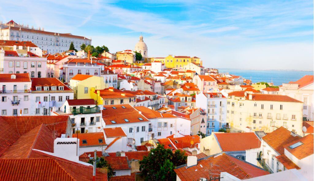 Vista panorâmica do bairro de Alfama, em Lisboa, Portugal, com as suas casas típicas de fachadas coloridas e telhados vermelhos. Em destaque, vê-se ao fundo a cúpula do Panteão Nacional e, ao longe, o rio Tejo sob um céu azul com algumas nuvens finas.