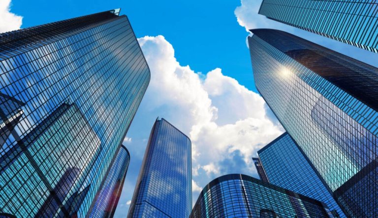 Fotografia tirada de baixo para cima mostrando arranha-céus de vidro espelhado sob um céu azul com nuvens brancas. A perspectiva acentua a altura e imponência dos edifícios, refletindo o céu nas suas fachadas modernas e reluzentes.
