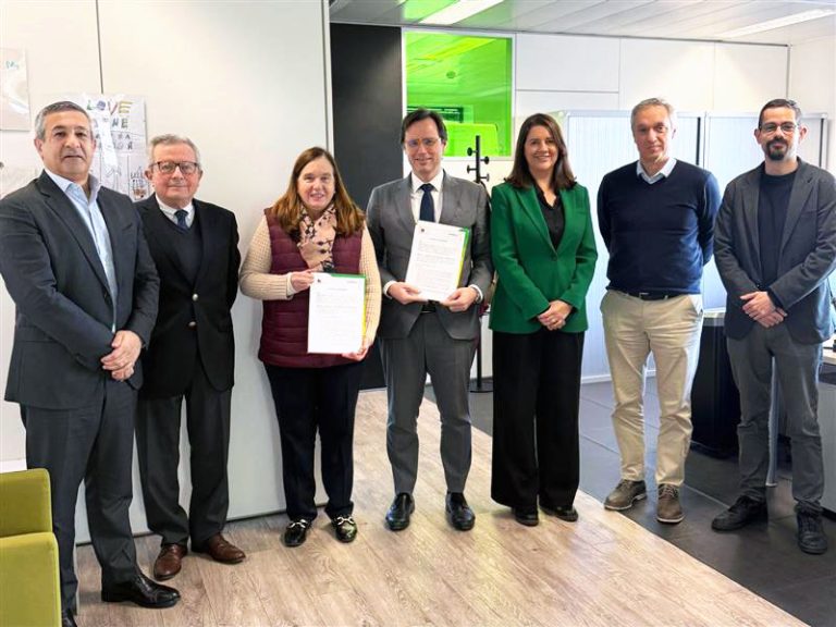 Sete pessoas estão alinhadas para uma fotografia em ambiente de escritório. Ao centro, duas delas seguram documentos oficiais. Todos sorriem e usam vestuário formal ou semi-formal. A imagem representa uma parceria ou assinatura de protocolo entre a ADENE e a ENTRAJUDA, sugerido pelo contexto corporativo.