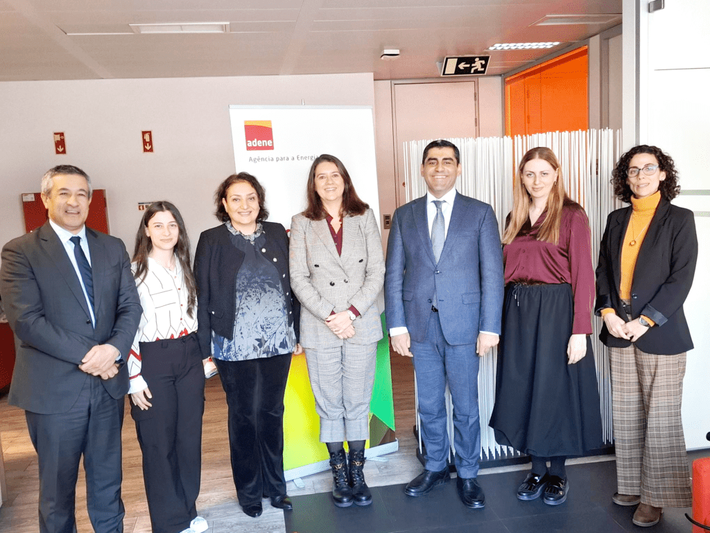 Sete pessoas posam para uma fotografia em ambiente corporativo, em frente a um expositor com o logótipo da ADENE – Agência para a Energia. O grupo é composto por quatro mulheres e três homens, todos vestidos com roupa formal. Estão sorridentes, lado a lado, e a fotografia parece registar um momento institucional ou de visita oficial nas instalações da ADENE.