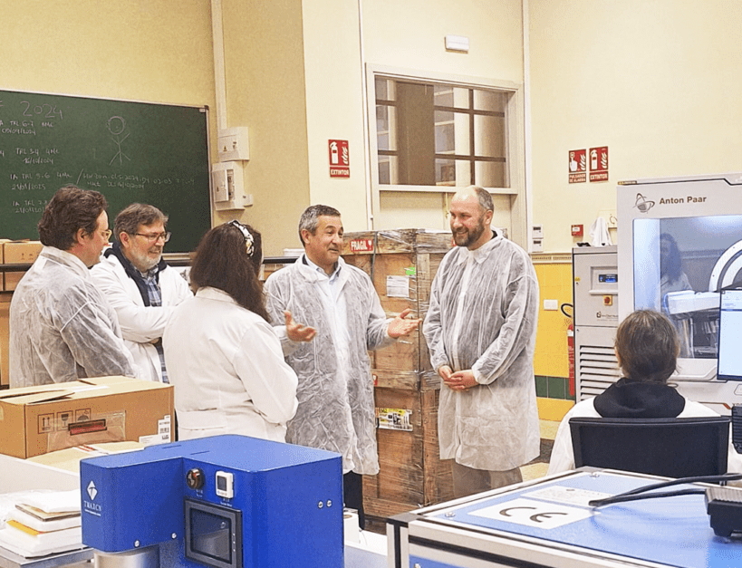 Um grupo de seis pessoas, quatro homens e duas mulheres, está reunido num laboratório. Todos usam batas ou vestuário protetor transparente. Estão a conversar informalmente junto a uma bancada com equipamentos laboratoriais de cor azul. Ao fundo, vê-se um quadro de giz com anotações, caixas empilhadas e um equipamento da marca Anton Paar. A cena transmite um ambiente de visita técnica ou demonstração.