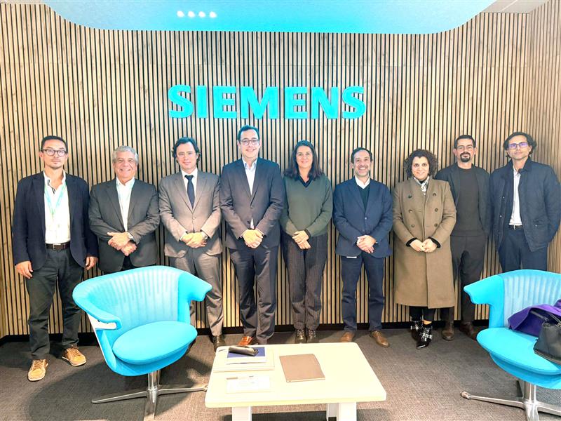 Fotografia de grupo com nove pessoas em traje profissional, alinhadas em frente a uma parede de madeira com o logótipo azul-turquesa da "SIEMENS" iluminado ao centro. Duas cadeiras azuis e uma mesa branca com documentos estão à frente do grupo, sugerindo uma reunião ou visita institucional.