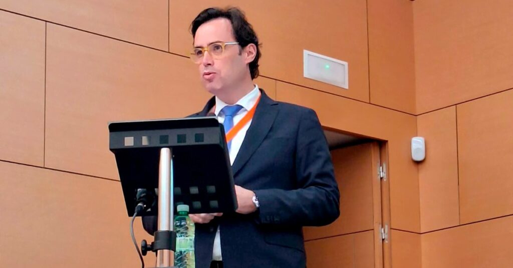 Fotografia do Nelson Lage, diretor da ADENE a discursar num púlpito durante um evento. Ele está vestido com fato escuro, camisa branca e gravata azul, usando um crachá laranja ao pescoço. Atrás dele, o fundo é composto por painéis de madeira clara. Uma garrafa de água está sobre o púlpito, que também contém um microfone.