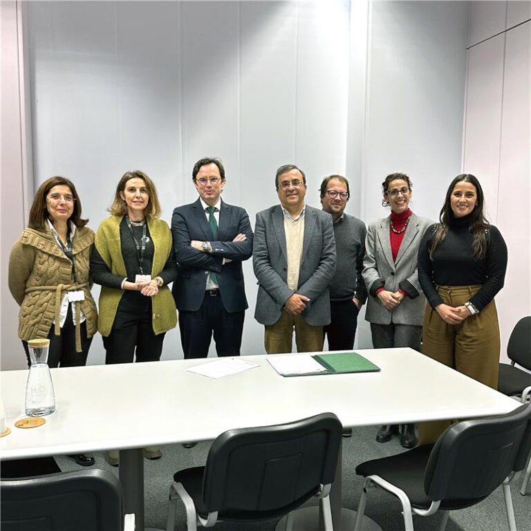 Um grupo de sete pessoas está posicionado lado a lado em frente a uma parede branca. Estão todos vestidos com roupas formais ou de trabalho. À frente do grupo há uma mesa branca com duas garrafas de vidro e alguns documentos pousados. Todos olham para a câmara com expressões neutras ou sorridentes.